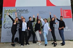 Na tle kilkumetrowej grafiki reklamowej grupa ośmiu osób stoi i uśmiecha się do zdjęcia. Na górze grafiki na czerwonym tle około 3 metrowe białe i czarne liczby 2025. Dół grafiki szary z mniejszymi czarnymi napisami.