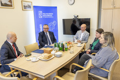 Na zdjęciu widać spotkanie w sali konferencyjnej Politechniki Rzeszowskiej. Przy stole siedzi pięć osób, na stole znajdują się napoje, filiżanki, ciastka i pudełko. W tle widoczny jest niebieski baner uczelni, telewizor z kamerą oraz zegar ścienny. 
