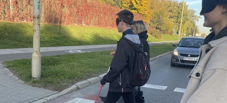 Na zdjęciu studenci przechodzący przez pasy dla przechodniów. Wyposażeni w laski dla niewidomych, opaski na oczy oraz ortezy. 