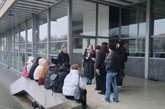 W podcieniu budynku ze szklanymi ścianami zgromadzona podczas rozmowy grupa kilkunastu osób. Część osób siedzi na betonowej ławce.