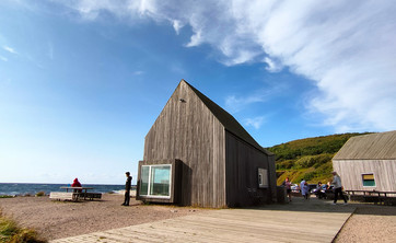 Zdjęcie: Pośrodku rozległej przestrzeni w naturalnym surowym krajobrazie zlokalizowany parterowy budynek. Bryła prosta z wysokim dachem, całość wykończona pionowym szarym deskowaniem. W tle zielone wzgórze i jaskrawo niebieskie niebo.