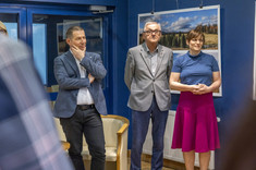 W pomieszczeniu stoją obok siebie trzy osoby, dwóch mężczyzn i kobieta po lewej stronie. Wszyscy zwróceni w jednym kierunku. W tle na granatowej ścianie zawieszone plansze z dużymi kolorowymi fotografiami.