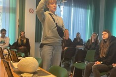 W pomieszczeniu na pierwszym planie stoi kobieta, w wysoko uniesionej ręce trzyma pracę plastyczną i tłumaczy coś zebranym za nią słuchaczom.