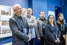 We wnętrzu z ciemnoniebieskimi ścianami, na tle plansz wywieszonych na ścianie, stoi grupa pięciu osób. Mężczyźni po lewej się uśmiechają.