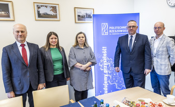 Na zdjęciu widoczna jest grupa osób stojących w pomieszczeniu biurowym. W tle znajduje się roll-up z logo Politechniki Rzeszowskiej im. Ignacego Łukasiewicza. Na stole przed grupą znajdują się materiały promocyjne.