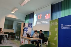 W lekko przyciemnionym pomieszczeniu po środku wysoko zawieszony jasny ekran, przed nim, przy stole siedzi dwóch prelegentów, mężczyzna i kobieta. W tle kilka wysokich na około 2 metry plansz w zielonym lub niebieskim kolorze z białymi napisami.