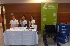 Na zdjęciu trzy uśmiechnięte studentki siedzą za stołem przykrytym białym obrusem, na którym znajdują się ulotki, okulary i gadżety. Za nimi stoi zielony baner Katedry Urbanistyki i Architektury oraz wózek inwalidzki. Obok widoczne są dwa niebieskie kostki z napisem IMMERSJA.