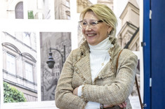 Kobieta ubrana na jasno stoi na tle planszy ze zdjęciami i się uśmiecha.