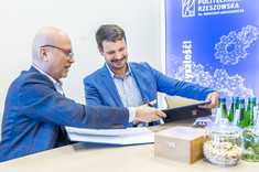 Dwóch mężczyzn siedzi przy stole i wymienia się podpisanymi umowami. W tle biała ściana z niebieskim pasem z napisem Politechnika Rzeszowska.