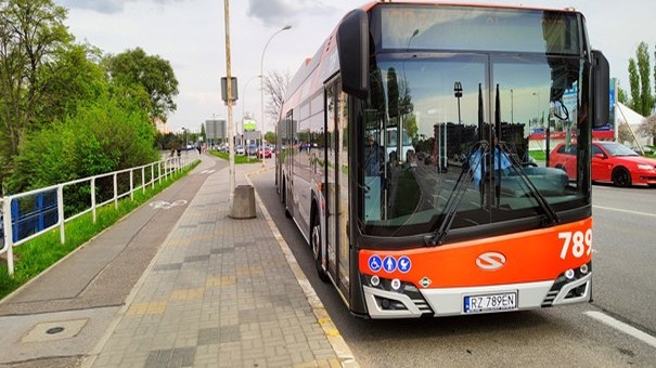 Na kolorowym zdjęciu po środku autobus stojący na przystanku. Po lewej stronie widoczny chodnik i ścieżka rowerowa, w tle zieleń.