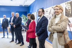 W jasno oświetlonym wnętrzu, z ciemnoniebieskimi ścianami, na tle plansz wywieszonych na ścianie stoi w rzędzie siedem elegancko ubranych osób.