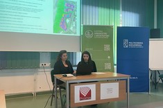 W lekko przyciemnionym pomieszczeniu po środku wysoko zawieszony jasny ekran, przed nim, przy stole siedzą dwie prelegentki. W tle kilka wysokich na około 2 metry plansz w zielonym lub niebieskim kolorze z białymi napisami.