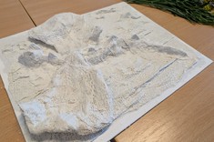 Na jasnym stole leży praca plastyczne. Na kwadratowej podstawie ukształtowane różnej wysokości wzniesienia. Praca jest cała biała.