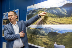 Na pierwszym planie stoi autor wystawy, mężczyzna w ciemnej marynarce, wskazuje ręką na fotografię. W tle na granatowej ścianie zawieszone plansze z dużymi kolorowymi fotografiami pejzaży górskich.