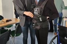 Zdjęcie wewnątrz sali. Na zdjęciu widoczne na głównym planie dwie osoby. Kobieta ubrana w symulator starości oraz mężczyzna pomagający jej go ubrać.