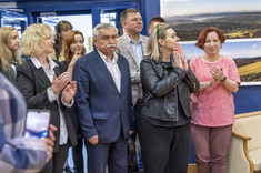 Ujęcie pomieszczenia, w którym stoi grupa kilkunastu osób. Wszyscy zwróceni w jednym kierunku, biją brawo i uśmiechają się. W tle na granatowej ścianie zawieszone plansze z dużymi kolorowymi fotografiami.