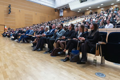 Zdjęcie sali wykładowej w całości wypełnionej słuchaczami. Miejsca siedzące są ustawione w równoległych rzędach, które stopniowo wznoszą się ku tyłowi pomieszczenia.