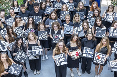 Zdjęcie wykonane z góry. Grupa około dwudziestu studentów i studentek ubranych na czarno spogląda w stronę obiektywu, każdy z nich w rękach trzyma biało-czarną makietę.