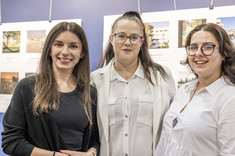 Trzy kobiety z ciemnymi długimi włosami, jedna ubrana na czarno, dwie na biało stoją obok siebie i uśmiechają się do zdjęcia. 