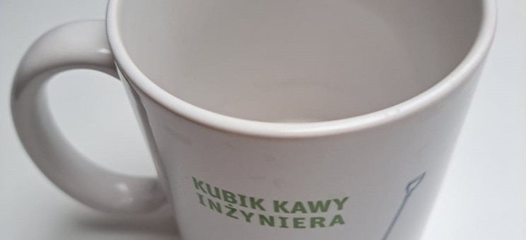 Zdjęcie przedstawia biały bubek z uchem. Na kubku namalowane granatowe grafiki łopata z napisem łyżeczka i wiadro z napisem woda. Nad nimi zielony napis KUBIK kawy inżyniera.