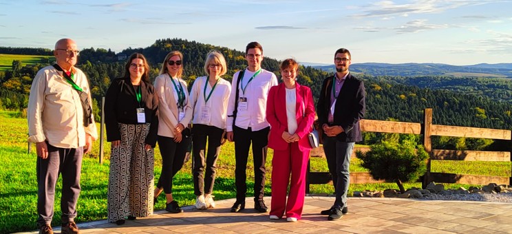 Grupa siedmiu osób pozuje do zdjęcia na tle naturalnego krajobrazu. Wszyscy są uśmiechnięci. W tle zielone wzgórza pokryte lasami i niebieskie niebo.