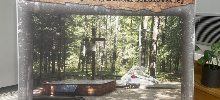 Na zdjęciu widoczny jest plakat przedstawiający miejsce pamięci w lesie turzańskim, nazwane „Mały Katyń Ziemi Sokołowskiej”. Centralnym punktem fotografii jest pomnik z krzyżem, otoczony drzewami. Na pomniku znajduje się tablica pamiątkowa, a przed nim widoczne są kwiaty i znicze.
