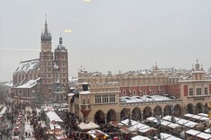 Widok z góry na rynek w Krakowie. Rynek w aurze zimowej wypełniony licznymi straganami na tle zabytkowego budynku Sukiennic z podcieniami i charakterystycznej sylwety Kościoła Mariackiego z dwiema wieżami o różnej wysokości.