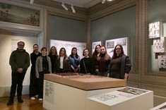 We wnętrzu z historycznymi zdobieniami i przyciemnionym światłem, na ścianach zawieszone plansze. Pośrodku za prostokątną bryłą grupa kilkunastu osób pozuje do zdjęcia.