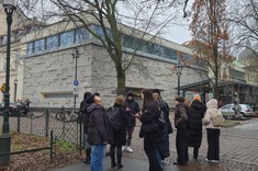 Pora jesienna. Zdjęcie na zielonym placu przed betonową bryłą budynku stoi grupa kilkunastu osób. Wokół liczne drzewa.