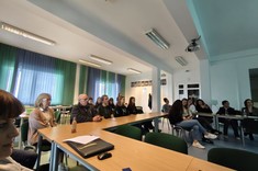 W lekko przyciemnionym pomieszczeniu za stołami ułożonymi w kształcie litery C siedzą słuchacze seminarium. 