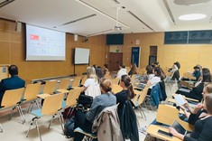 W jasnym wnętrzu sali wykładowej ze ścianami z drewnopodobną wykładziną i z białym sufitem zgromadzona kilkudziesięcioosobowa grupa słuchaczy. W tle jasny ekran z wyświetloną prezentacją. 