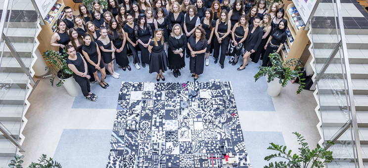 Zdjęcie wykonane z góry. W centrum ujęcia na podłodze ułożony kwadrat około 3 x 3 metry z biało-czarnych kwadratowych makiet. Wokół zgromadzona grupa kilkudziesięciu osób. wszyscy ubrani na czarno. 