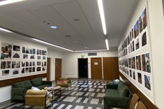 W pomieszczeniu z jasnymi ścianami i sufitem na ścianach po prawej i lewej stronie zawieszone duże plansze z wyeksponowanymi fotografiami w stonowanych barwach.