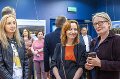 Na pierwszym planie trzy kobiety stoją obok siebie, uśmiechają się do fotografii.  W tle kilkanaście osób na tle ciemnej ściany z prezentowanymi zdjęciami.