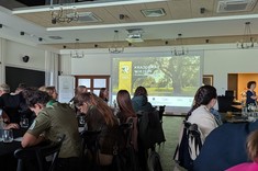 W jasnym pomieszczeniu liczne osoby siedzące przy równolegle ustawionych stołach. W tle na ścianie wyświetlona prezentacja.