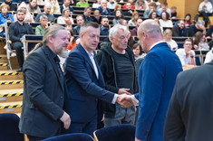 Czterech mężczyzn podczas oficjalnego przywitania. Trzech zwróconych przodem do zdjęcia. W tle uczestnicy wykładu.