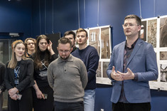 We wnętrzu z ciemnoniebieskimi ścianami, na tle plansz wywieszonych na ścianie, stoi grupa kilku osób, a mężczyzna po prawej przemawia.