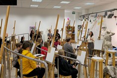 Widok na salę plastyczną. Około 30 uczestników stojących przy sztalugach – drewnianych trójnogach słucha prowadzącej.