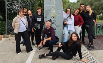 Grupa dziecięciu osób, swobodnie rozmieszczonych wokół pionowego banneru reklamowego pozuje do wspólnego zdjęcia. Wszyscy się uśmiechają. Na banerze, na szarym tle umieszczony napis w pionie Artificial Collective i czerwony znak biennale.  W tle wysokie drzewo.