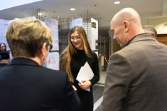 Na zdjęciu młoda kobieta ubrana w czarną sukienkę odbiera nagrodę. Na perwszym planie dwie osoby stojące plecami do fotografa. W tle wystawa: plansze zawieszone na metalowych siatkach,