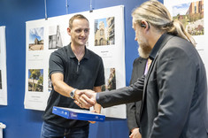 Mężczyzna w ciemnej marynarce wręcza nagrodę mężczyźnie w ciemnej koszulce.  W tle plansze ze zdjęciami.