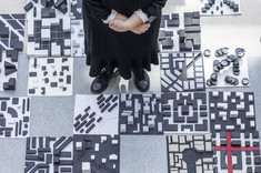 Zdjęcie wykonane z góry. Pośrodku stoi kobieta ubrana na czarno, spogląda w stronę obiektywu. Wokół na podłodze rozłożone biało czarne makiety. .