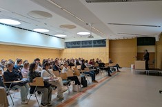 Wewnątrz sali z jasnym drewnopodobnym wykończeniem ścian i białym sufitem zgromadzonych jest kilkadziesiąt osób podczas wykładu. Na zdjęciu z prawej strony stoi prelegent na niewielkim podwyższeniu. Na pierwszym planie siedzący słuchacze. 