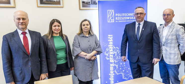 Na zdjęciu widoczna jest grupa osób stojących w pomieszczeniu biurowym. W tle znajduje się roll-up z logo Politechniki Rzeszowskiej im. Ignacego Łukasiewicza. Na stole przed grupą znajdują się materiały promocyjne. 