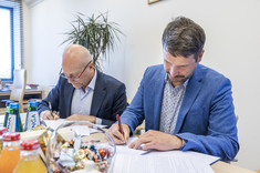 Dwóch mężczyzn elegancko ubranych siedzi przy stole i podpisuje dokumenty.