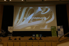 Na zdjęciu widzimy scenę w nowoczesnej sali audytoryjnej. Na dużym ekranie wyświetlony jest tytuł prezentacji. Tłem slajdu jest futurystyczne zdjęcie wnętrza budynku o nowoczesnych, organicznych kształtach. 