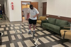 Mężczyzna w białej koszulce i czarnych szortach idzie po korytarzu w czarnych okularach symulujących wadę wzroku. Używa laski do nawigacji po ścieżce z taśmy ułożonej na podłodze. W tle widać zieloną sofę, fotele i otwarte drzwi do sali wykładowej.