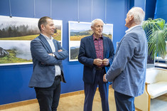 Trzech mężczyzn, ubranych w ciemno niebieskie garnitury stoi obok siebie i rozmawia. Uśmiechają się do siebie. W tle na granatowej ścianie zawieszone plansze z dużymi kolorowymi fotografiami.