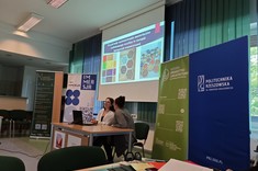 W lekko przyciemnionym pomieszczeniu po środku wysoko zawieszony jasny ekran, przed nim, przy stole siedzą dwie prelegentki. W tle kilka wysokich na około 2 metry plansz w zielonym lub niebieskim kolorze z białymi napisami.