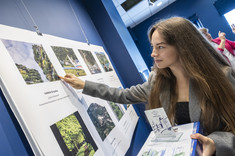 Zbliżenie na młoda kobietę z długimi włosami. Kobieta wskazuje ręką na zdjęcie, w drugiej ręce trzyma nagrodę.